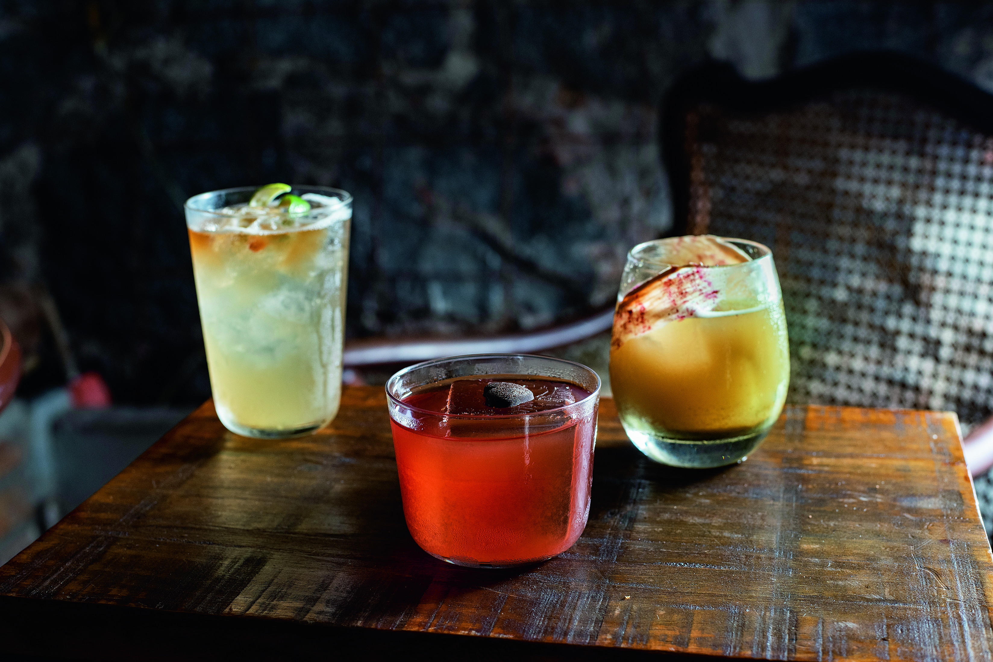 Tr&ecirc;s drinques sobre mesa de madeira. &Agrave; esquerda, copo alto com l&iacute;quido de cor clara, ao centro copo baixo com bebida avermelhada coberta por cora&ccedil;&atilde;o de chocolate e &agrave; direita, copo arredondado com drinque amarelado.