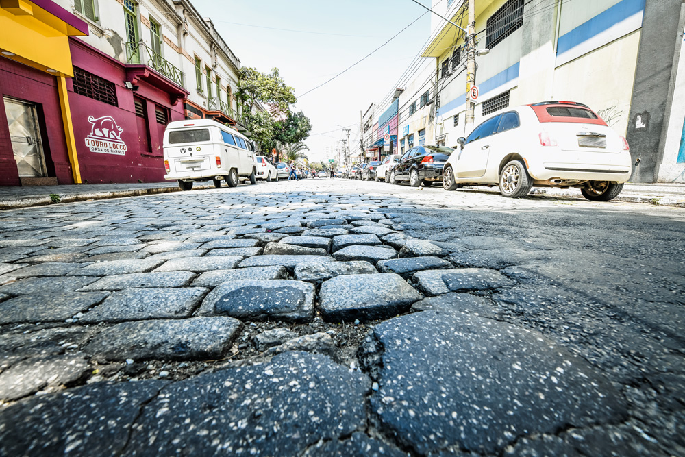 A imagem mostra uma foto bem próxima de um asfalto de paralelepípedos. No centro, há um buraco na rua.