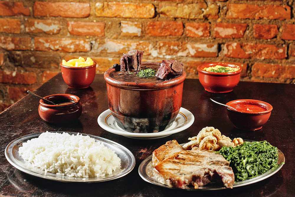 Feijoada servida em cumbuca no bar Veloso juntamente de uma porção de arroz, filés de bisteca e demais guarnições