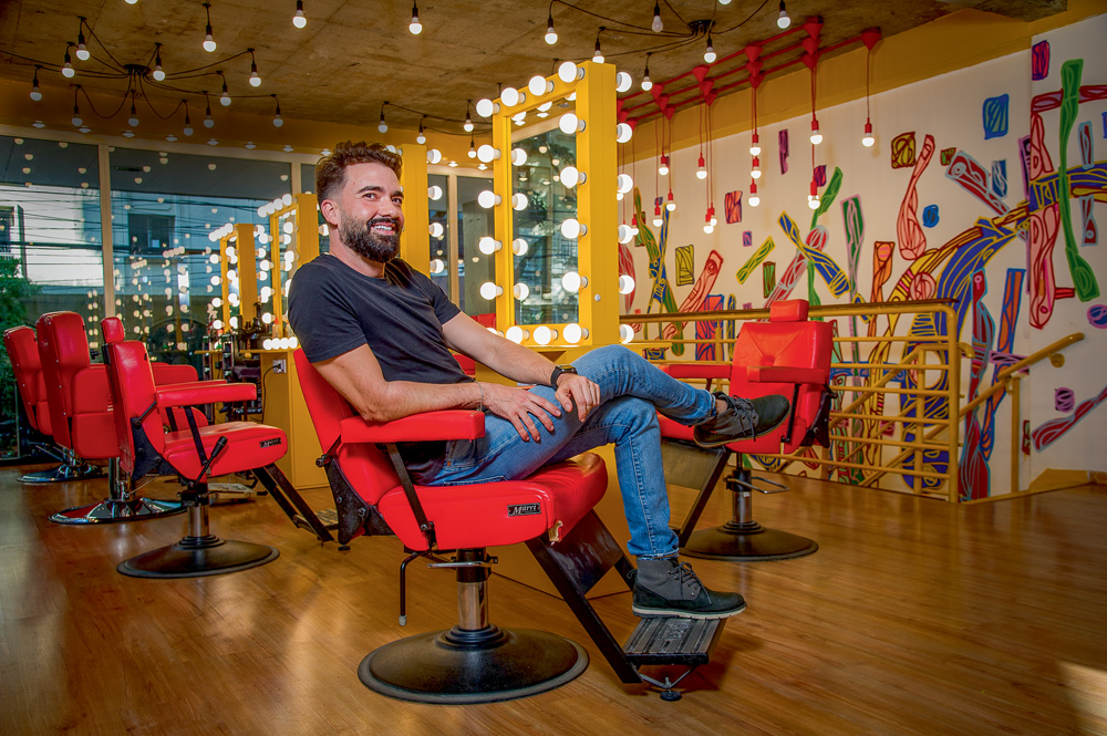 Felipe Ribeiro veste calças jeans e camiseta azul. Aparece sentado em cadeira vermelha de barbearia com as pernas cruzadas, sorrindo. Ao fundo, parede com grafites e espelhos iluminados.