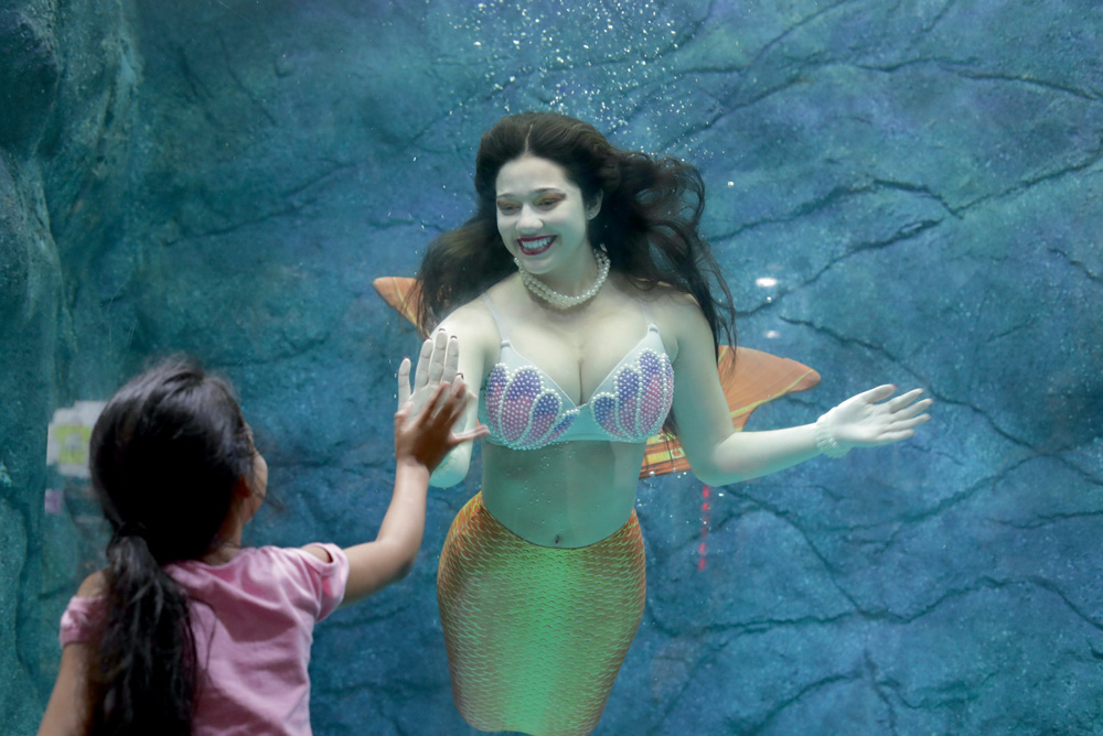 Uma mulher interpretando uma sereia em um tanque de água acena e sorri para uma criança que está do lado de fora do tanque