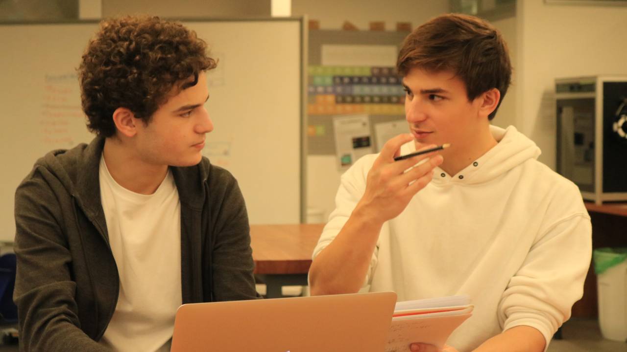 Dois jovens brancos sentados lado a lado conversam em frente a um computador da Apple. Um veste camiseta branca e uma blusa escura. Outro veste um moletom com capuz branco.