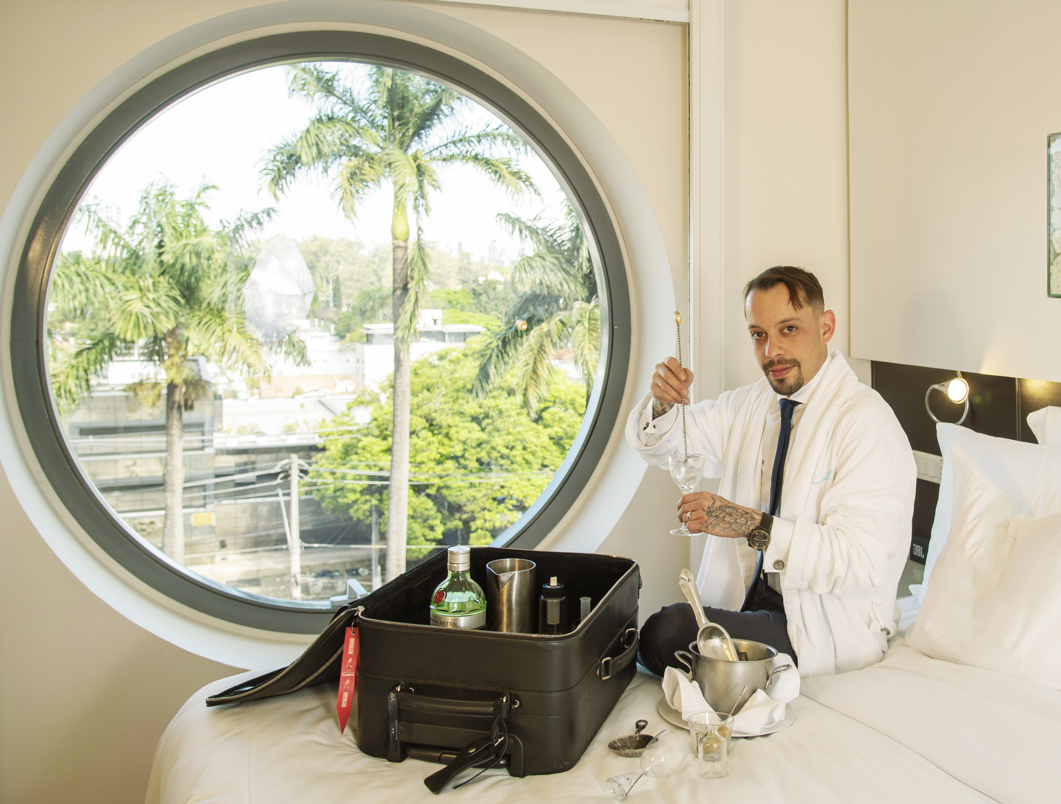 Gabriel Santana sentado em cama de hotel fazendo drinque com uma mala preta aberta com itens de preparo dentro.