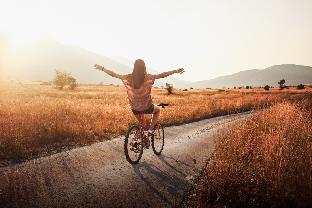 Uma jovem morena anda de bicicleta com os braços abertos, iluminada pelo pôr do sol de outono. Ela está em um caminho onde só há natureza