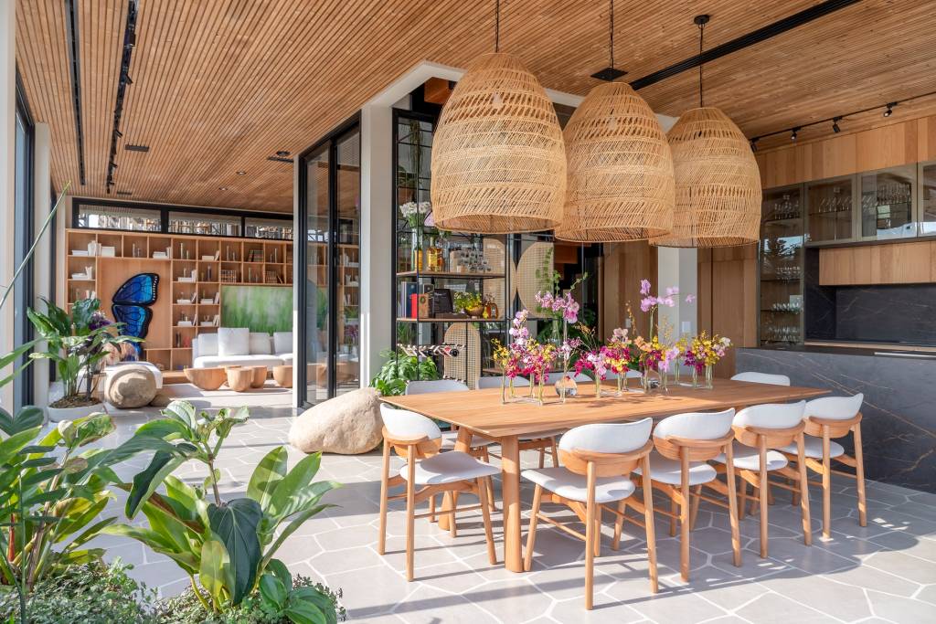 Residencial The Golf Village, na Riviera de São Lourenço, com espaço de almoço exibe grande mesa, plantas, lustres na cor de palha e vidraças ao fundo.