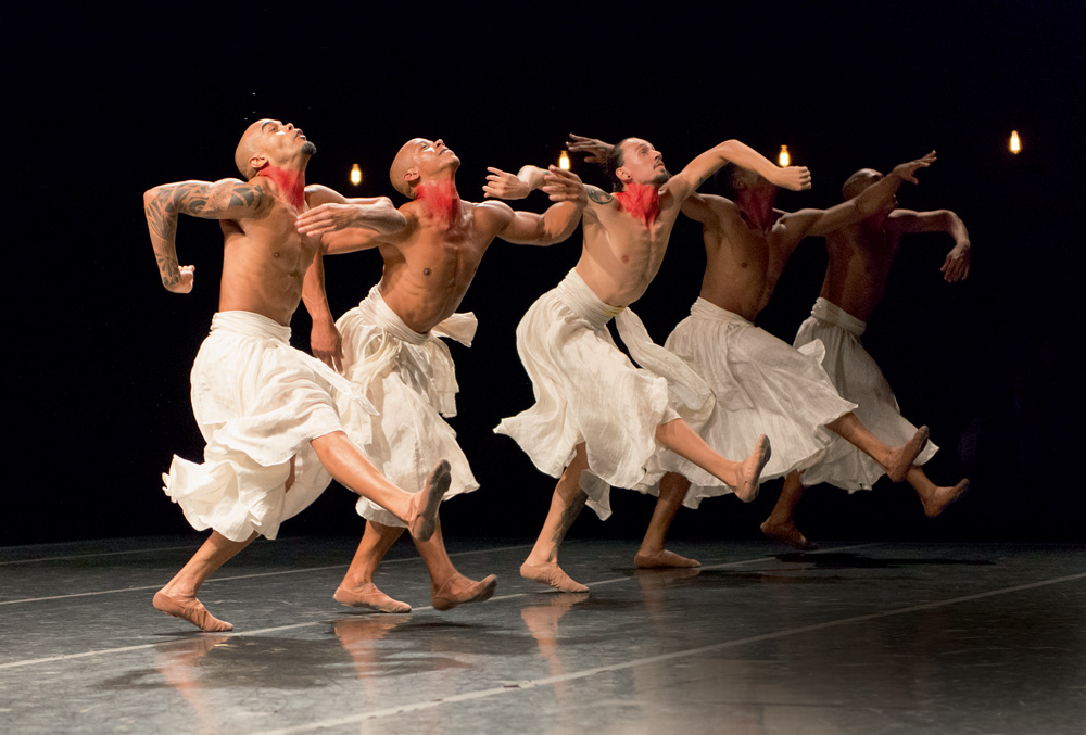A foto mostra cinco homens sem camisa, com uma grande saia branca e os pescoços marcados com tinta vermelha. Ele estão com os braços abertos e olhando para cima em cima do palco, durante a peça.