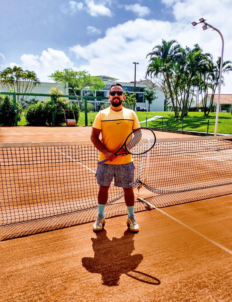 A imagem mostra Guilherme, já adulto, em uma quadra de tênis, segurando uma raquete de pé em frente a rede. Ele está de óculos escuro.