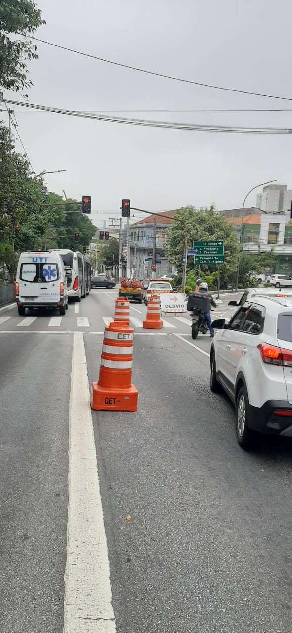 A imagem mostra faixas de circulação de veículos fechadas com cones laranjas e uma fila de carros se formando na parte permitida da rua.