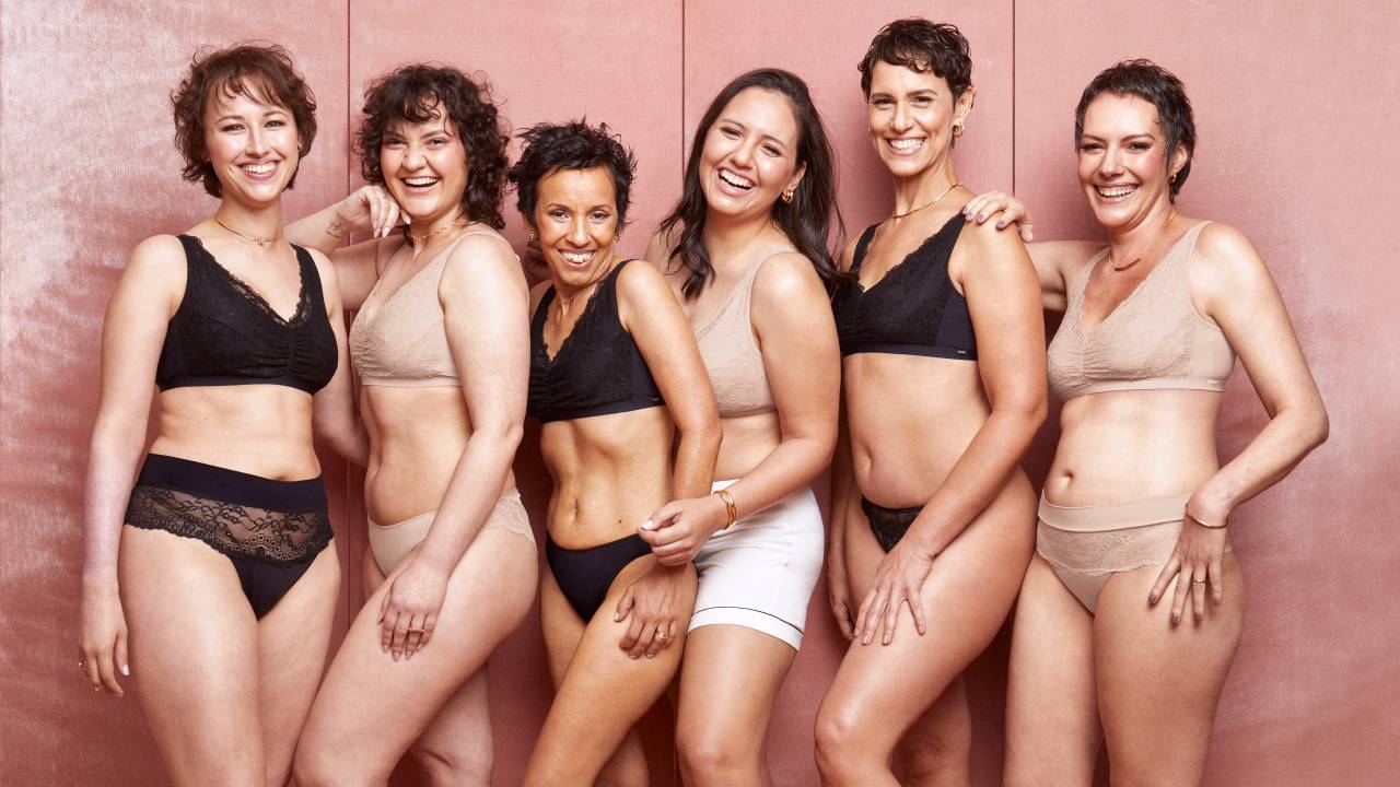Imagem mostra várias mulheres posando lado a lado de calcinha e sutiã, alguns na cor bege e outros na cor preta. Elas aparecem em um local com fundo rosa.