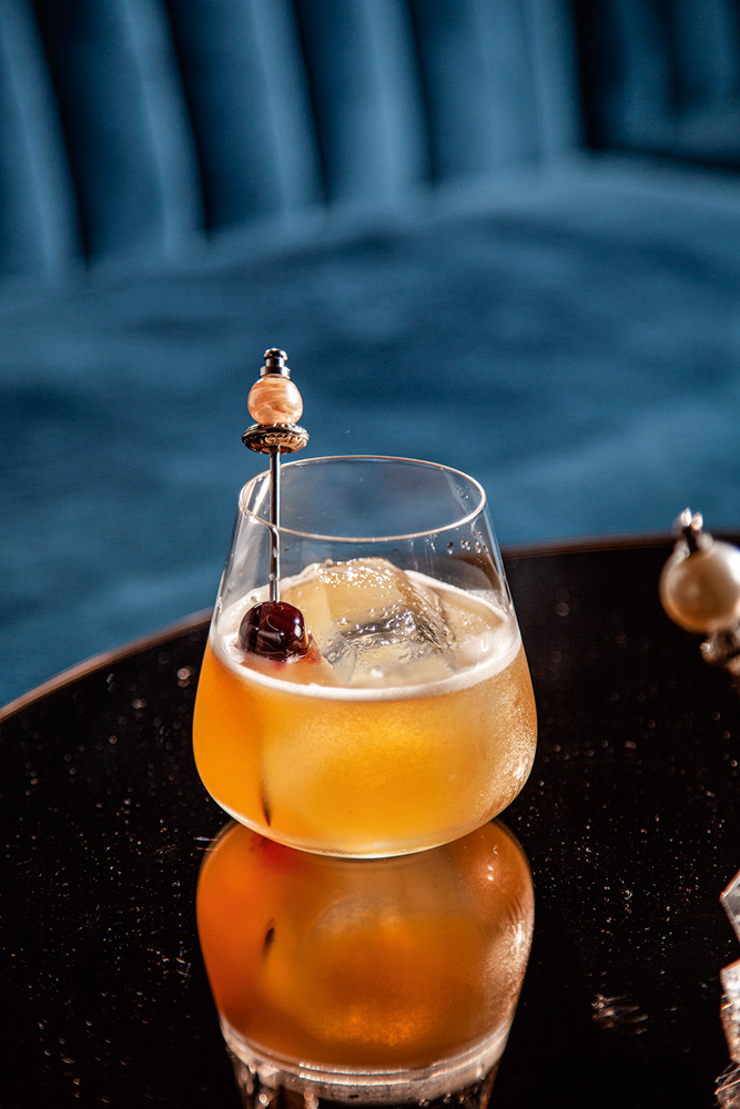 Drinque de cor alaranjada e decorado por uma cereja servido em um copo de vidro de fundo redondo