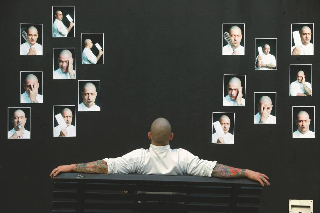 De costas, sentado em um banco de madeira pintado na cor preta, Henrique Fogaça encara fotos de si mesmo