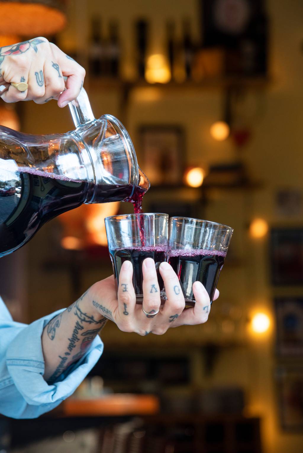 Ao centro, mão tatuada segurando dois copos de vidro sendo servidos de vinho em jarra à esquerda.