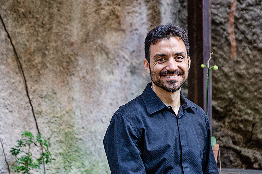 Um homem de camisa social preta posa sorrindo para a foto, no que parece ser um quintal
