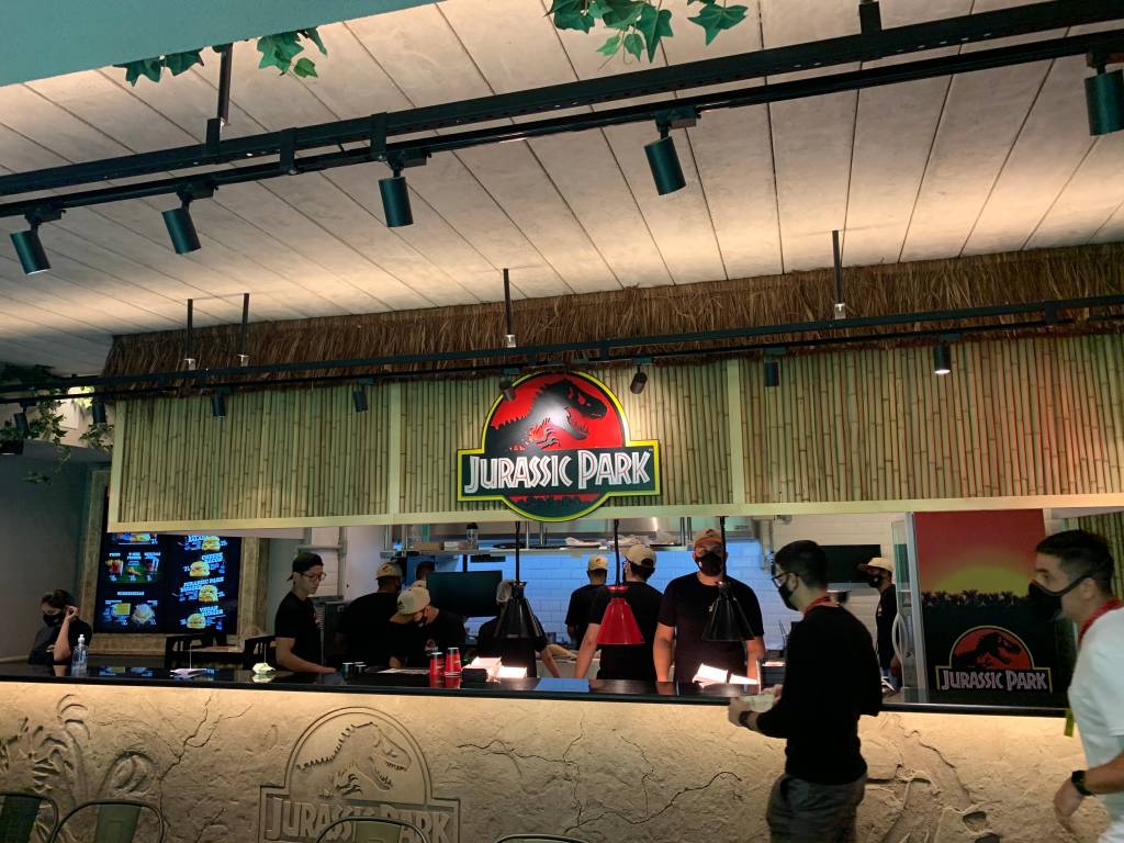 Cozinha da lanchonete Jurassic Park Burger Restaurant aberta para o público. Com balcão de cimento com dinossauro desenhado e teto de bambu.