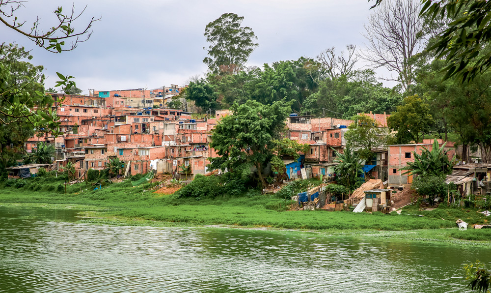 Na foto, uma represa com água verde e, ao fundo, uma favela. Entre a represa e a favela há um gramado