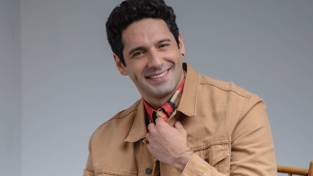 O ator João Baldasserini sorri para a câmera com uma mão na gola da camisa listrada. Por cima, veste uma jaqueta bege.