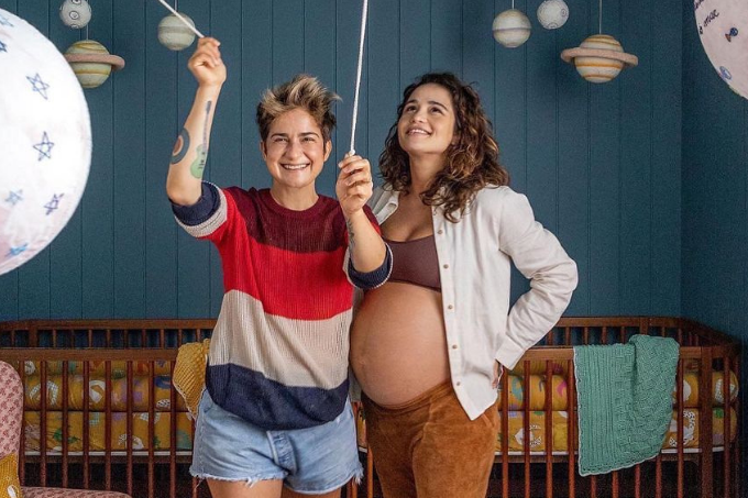 Nanda Costa e Lan Lanh posam em quarto de bebê, Nanda à direita com barriga de grávida aparente.