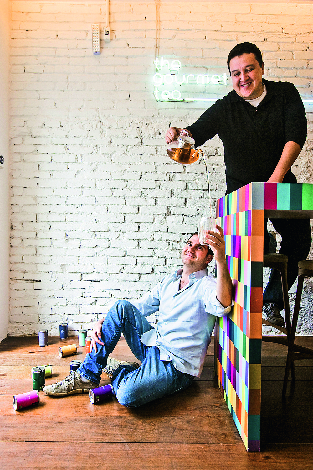 Um homem sentado no ch&atilde;o apoiado em uma mesa enquanto outro que est&aacute; na mesa serve de l&aacute; a x&iacute;cara de ch&aacute; do que est&aacute; no ch&atilde;o.