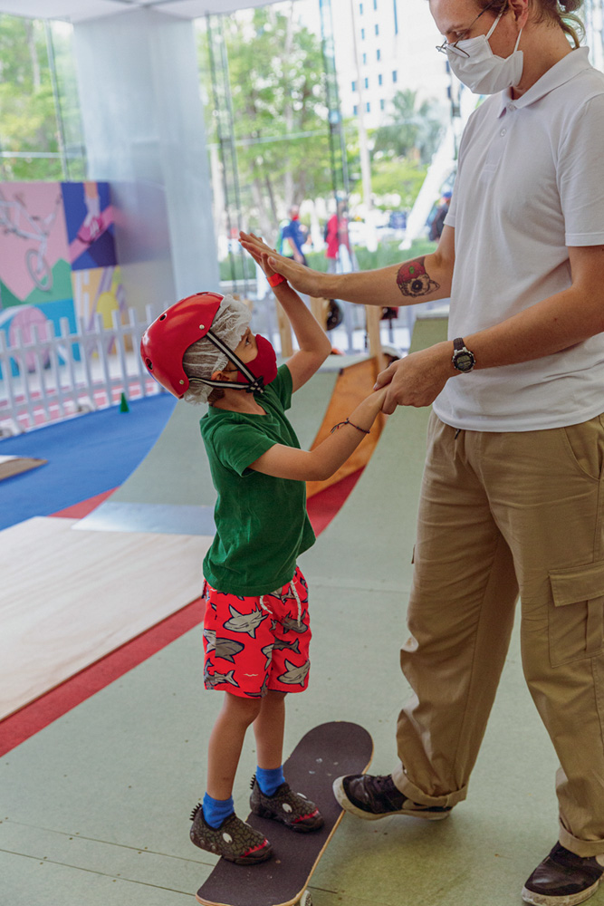 Uma criança, equipada com capacete, está em cima de um skate e bate na mão de um adulto ao lado dele.