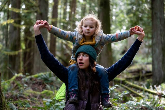 A imagem mostra uma jovem com sua filha em seus ombros, em uma floresta