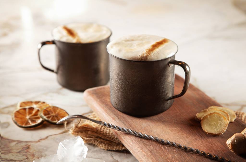 Duas canecas de moscow mule, com espuma por cima, sobre madeira ao lado de colher bailarina.