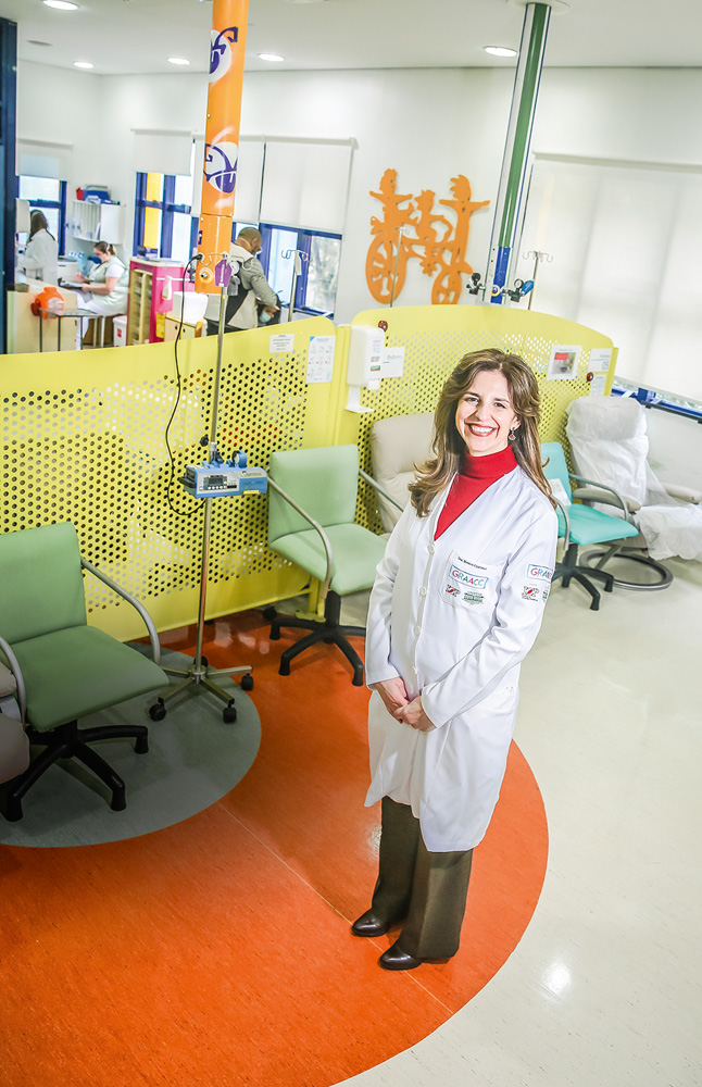 A foto mostra Monica Cypriano, de jaleco, em uma das salas do GRAAC. Ela está de pé, sorrindo para a câmera.