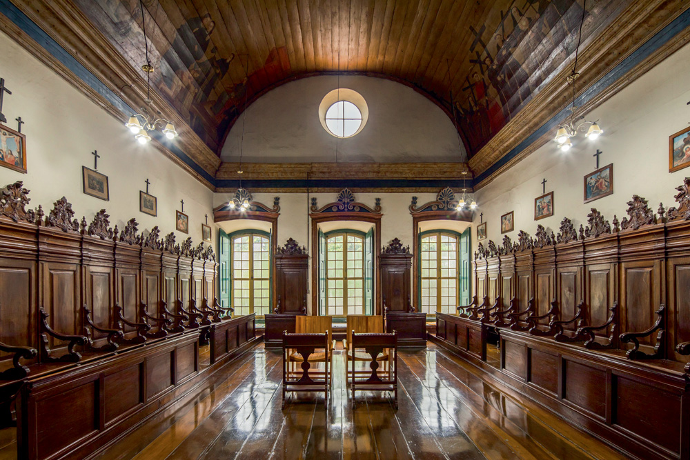sala de coro das freiras do mosteiro, com bastante madeira na constituição, no chão e nos bancos, que ficam em paredes opostas. no centro, poucas cadeiras, diferenciadas
