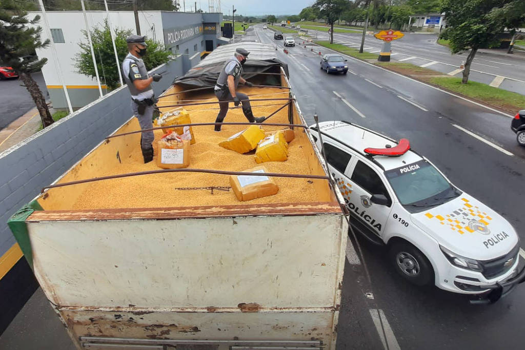 Policiais em cima de caminhão vistoriam veículo. Ao lado, carro de polícia.