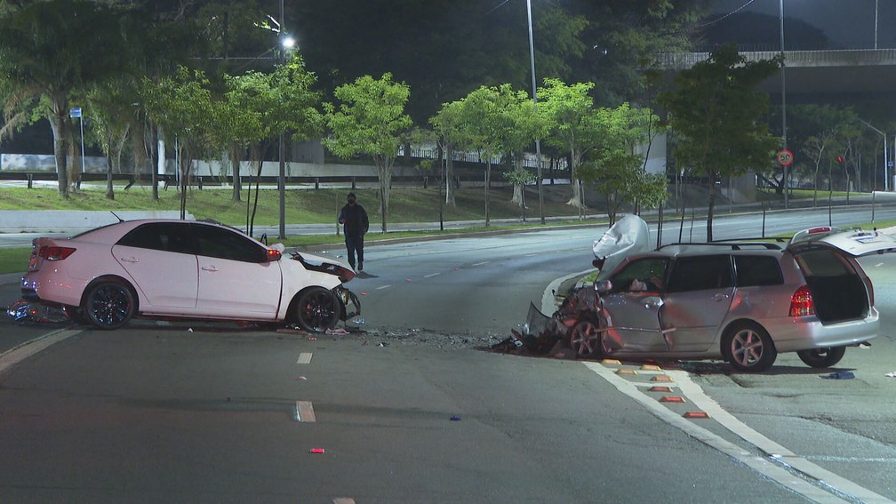 Imagem mostra dois carros parados após colisão em avenida. Ao fundo, uma silhueta de homem.