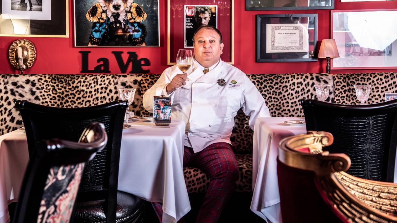 O chef Erick Jacquin sentado entre duas mesas no restaurante Président vestido de dólmã, segurando uma taça de vinho.