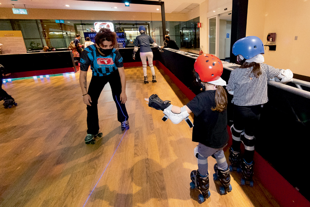 Um monitor auxilia duas crianças a brincarem em uma pista de patinação. Elas estão segurando nas bordas e ele no centro, na frente