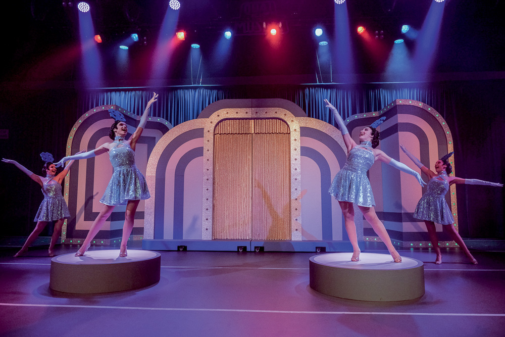 A foto mostra quatro bailarinas em dançando em frente a uma grande porta dourado no espetáculo Silvio Santos Vem Ai.