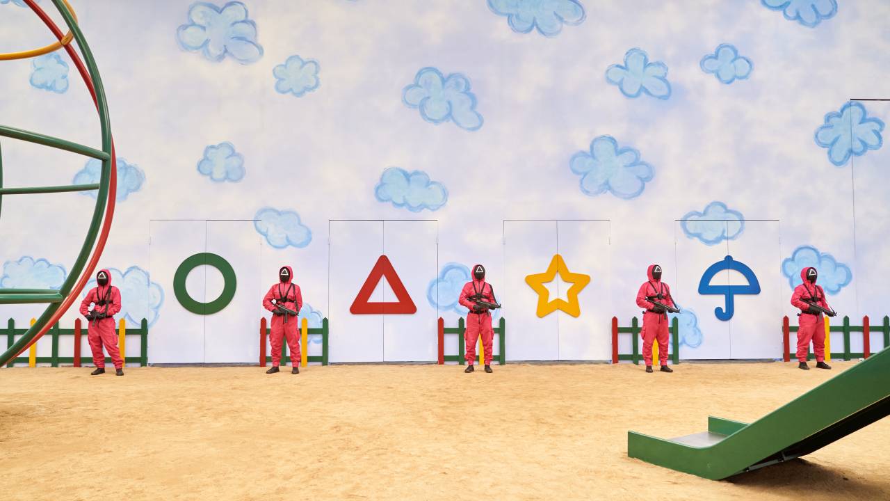 A imagem mostra uma cena da série Round 6, com pessoas mascaradas e com uniformes vermelhos posicionadas lado a lado