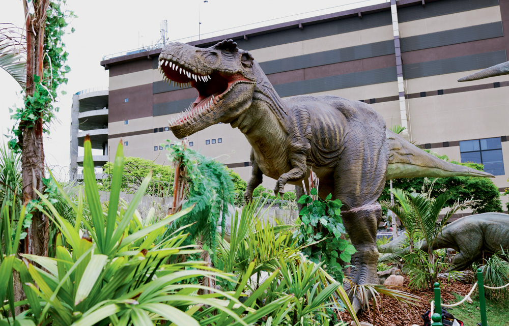 No estacionamento de um shopping, há plantas, e um gramado e, no centro, um boneco de dez metros de um dinossauro