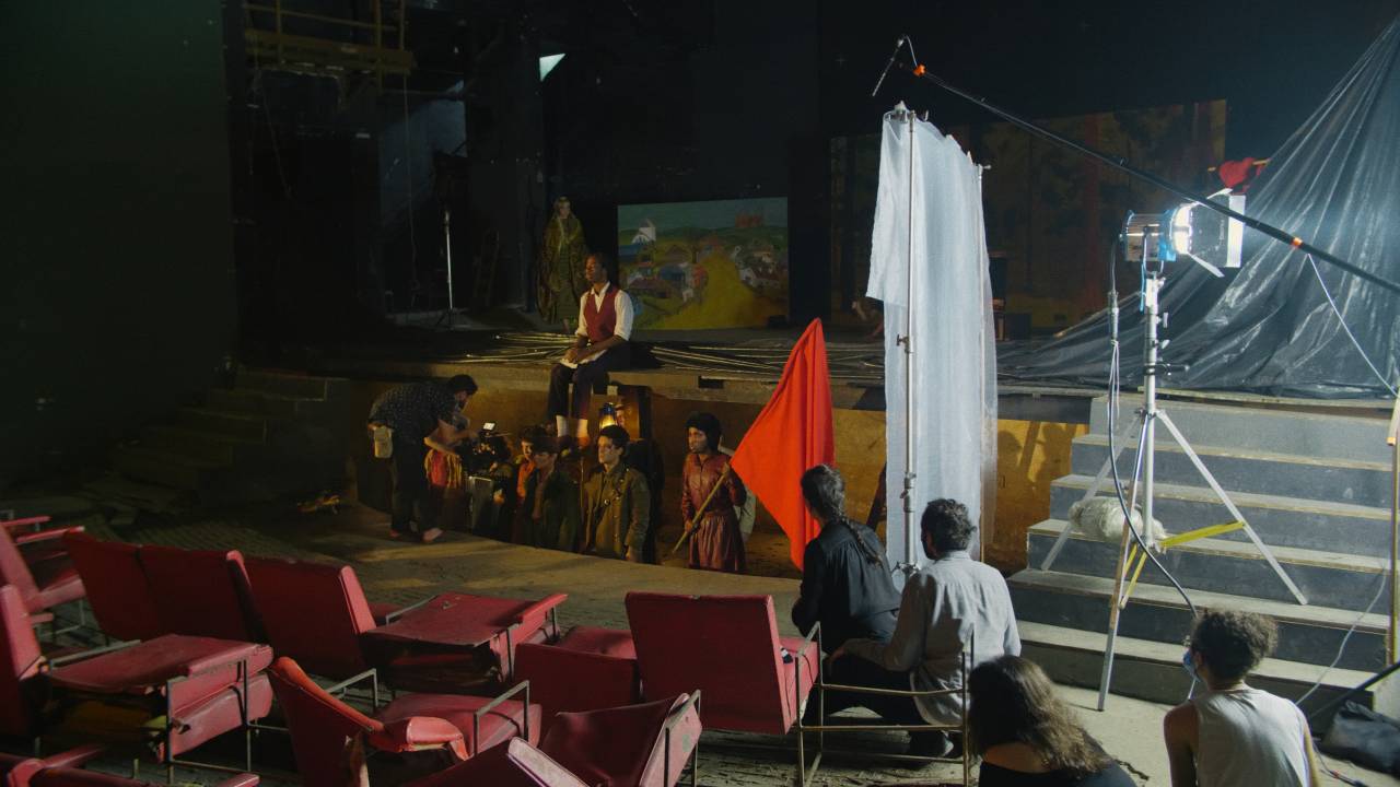 Imagem mostra cinegrafistas filmando grupo de pessoas com roupa de época que portam bandeiras vermelhas na bera de palco de teatro
