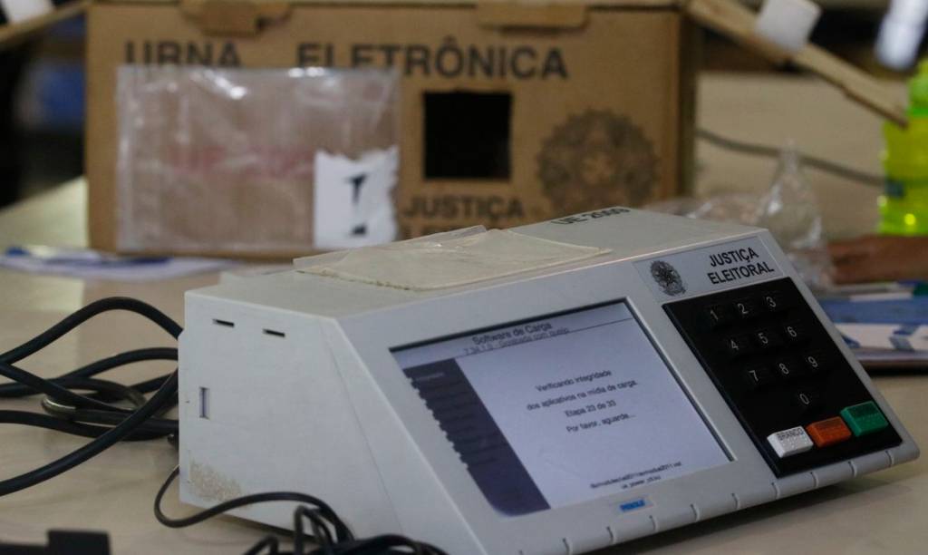 Imagem exibe urna eleitoral e caixa de papelão ao fundo.