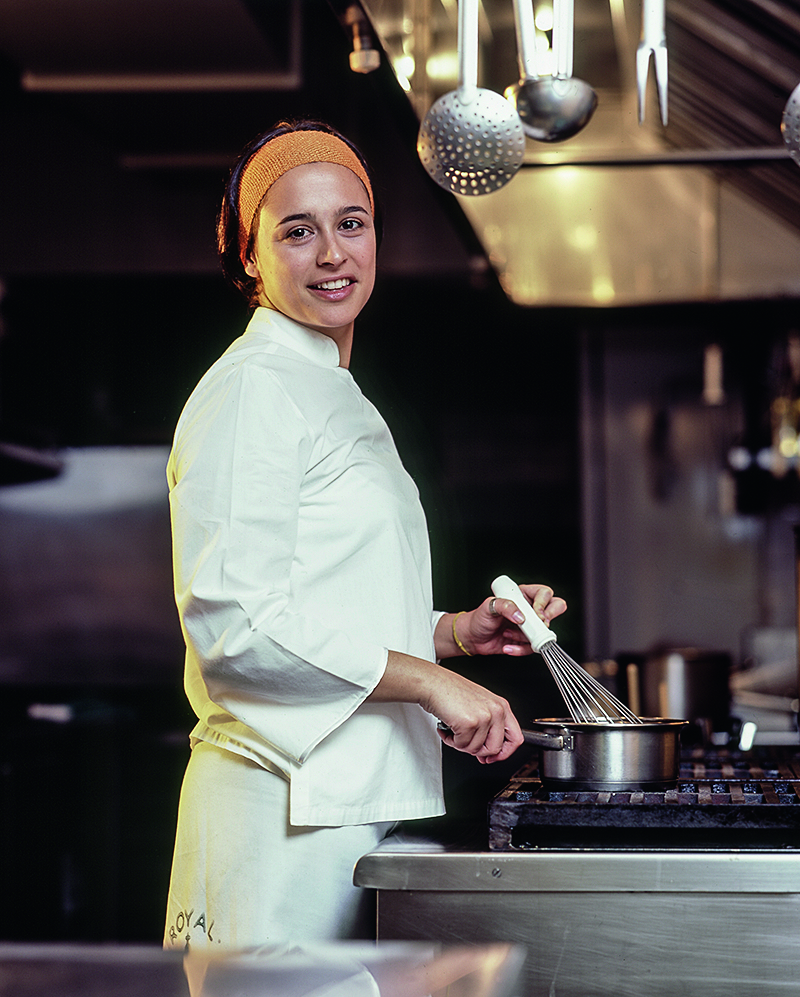 A chef Bel Coelho na cozinha do restaurante misturando uma panela no fog&atilde;o com um fouet.