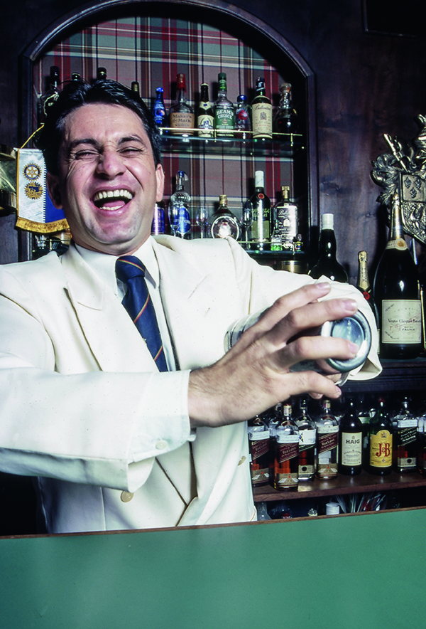 O bartender Deusdete Neres de Souza de terno branco e gravata azul batendo uma coqueteleira no atrás do balcão do bar Bistrô.