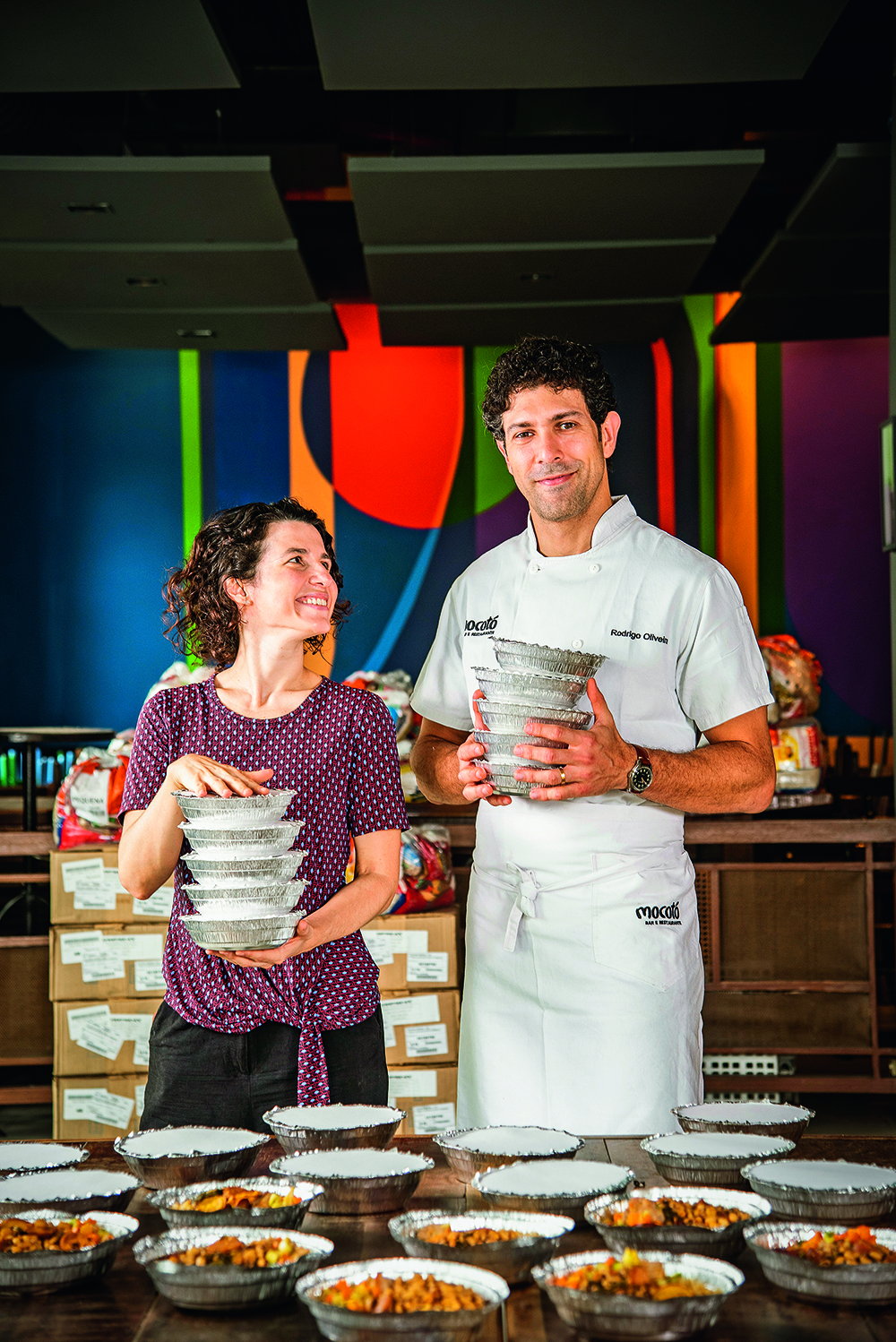 O chef Rodrigo Oliveira segurando quentinhas ao lado de sua esposa Adriana Salay, também segurando embalagens.