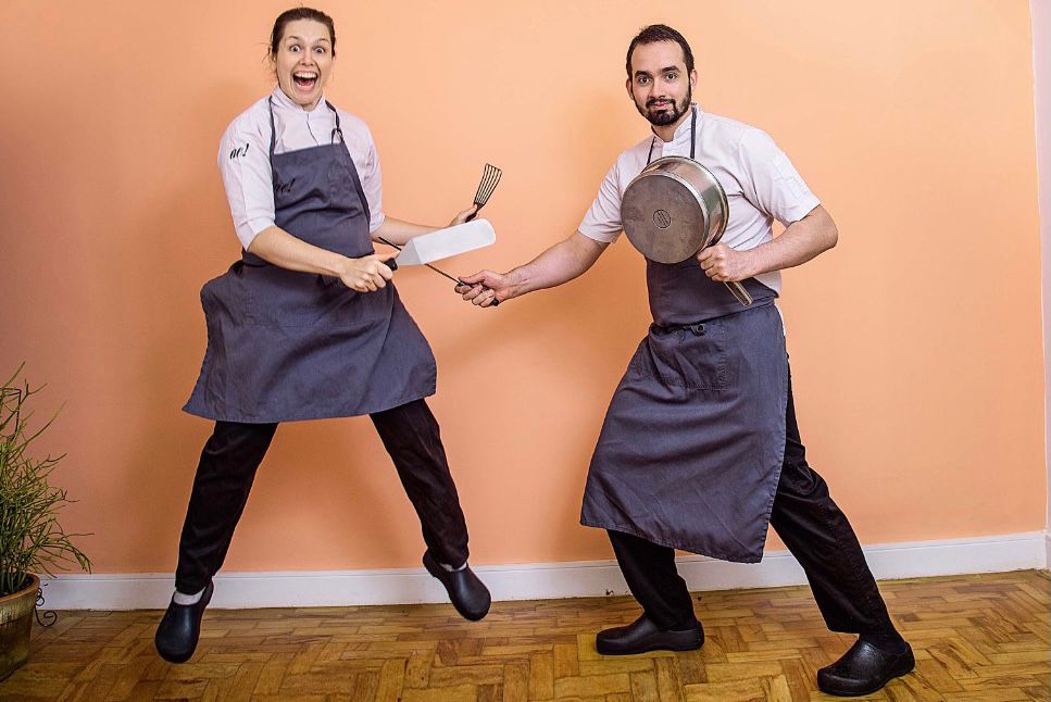 Chefs posam saltitantes em frente a uma parede de cor salmão enquanto segurando utensílios de cozinha