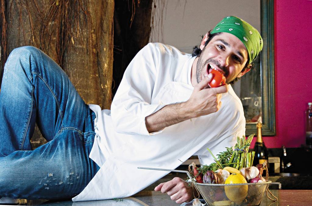 William Ribeiro posa sobre o balcão da cozinha enquanto segura um tomate próximo de sua boca