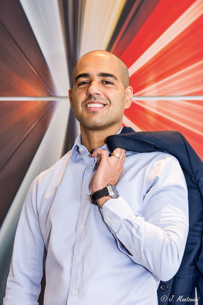 Um homem de camisa social azul e careca posa sorrindo para a foto. Ele segura um casaco com a mão apoiado nos ombros