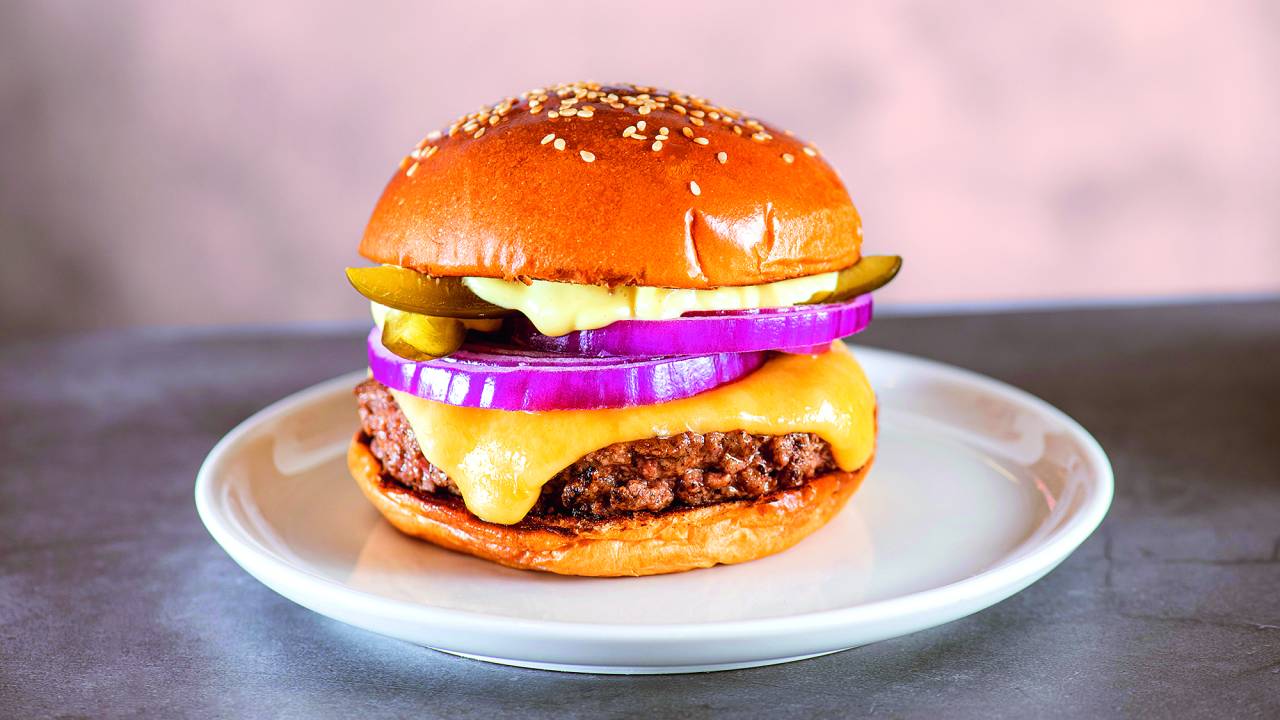 Hambúrguer sobre prato de louça branca com rodelas de cebola-roxa, queijo escorrendo pela carne no pão com gergelim.