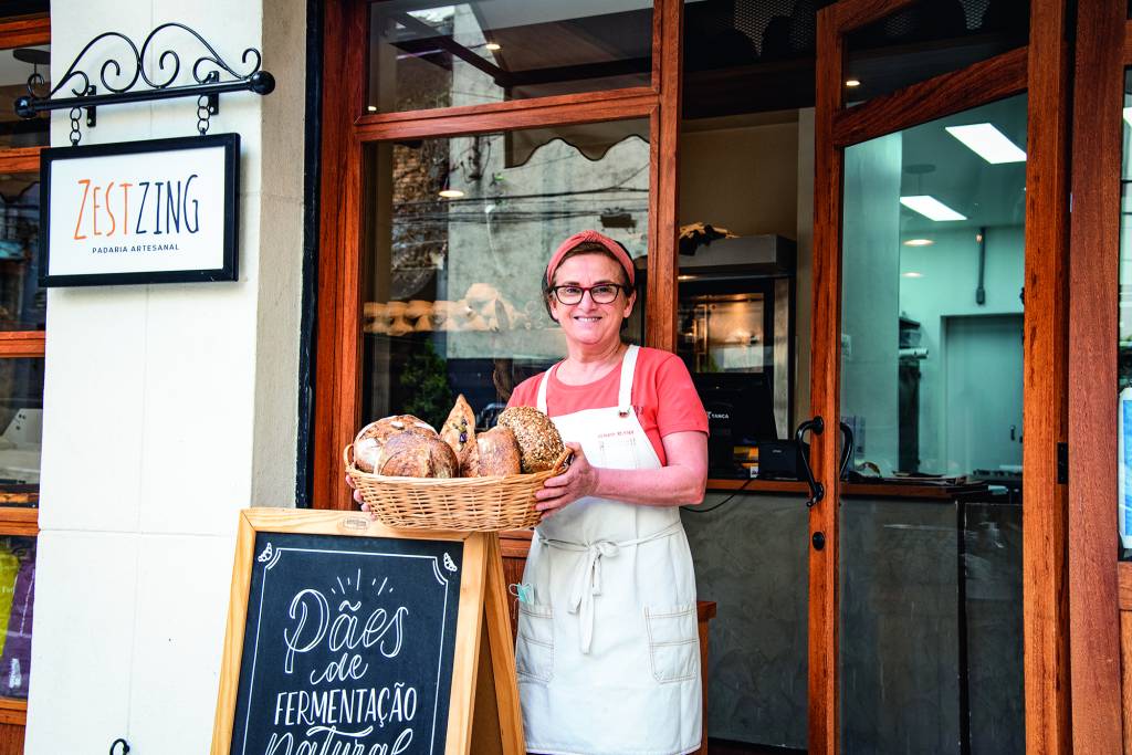 Claudia Rezende segurando cesta de pães sobre placa em frente à Zestzing Padaria Artesanal.