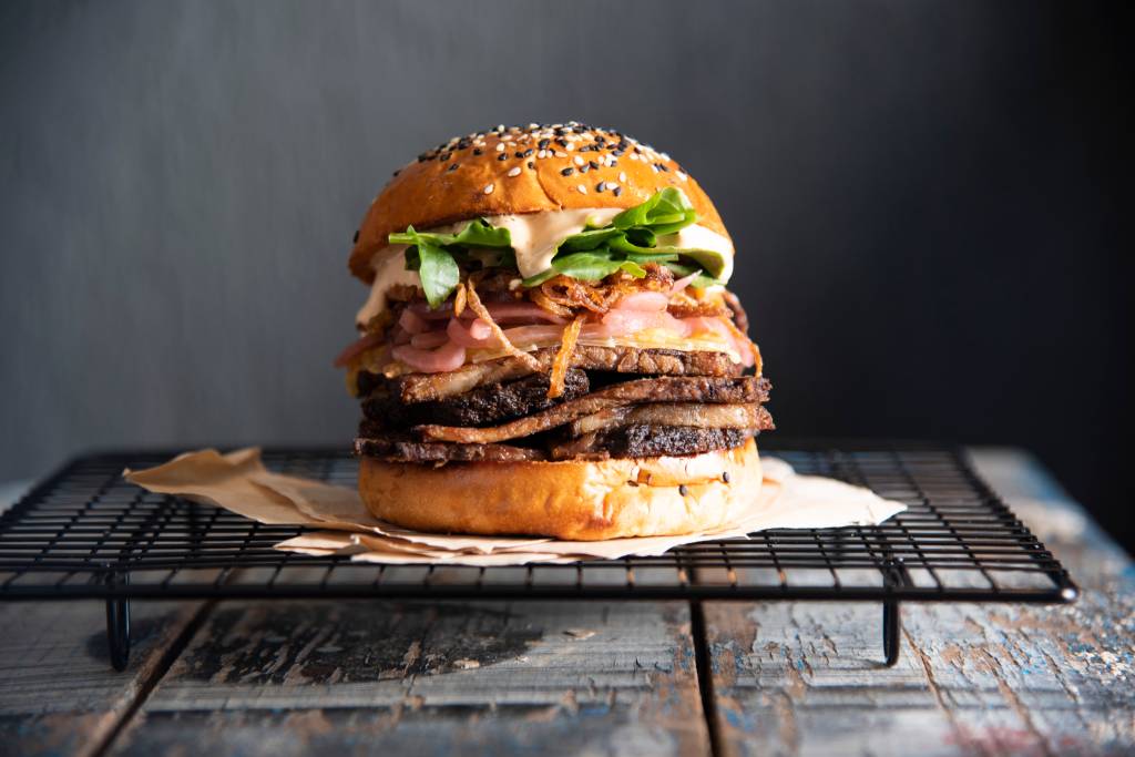 Sanduíche alto no pão de hambúrguer com camadas de brisket, cebola, molho e alface sobre suporte metálico preto em fundo cinza.