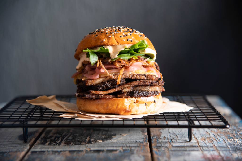 COMER & BEBER 2021 – Zoe Sandwich Shop Sanduíche alto no pão de hambúrguer com camadas de brisket, cebola, molho e alface sobre suporte metálico preto em fundo cinza.