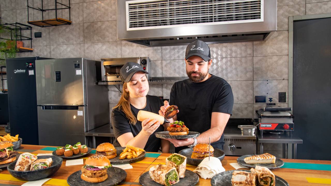 O casal de chefs Giovanna Perrone e Rodrigo Aguiar finalizando sanduíche juntos em cozinha aberta da Zoe Sandwich Shop com vários pratos com sandubas em frente à eles em balcão de madeira.