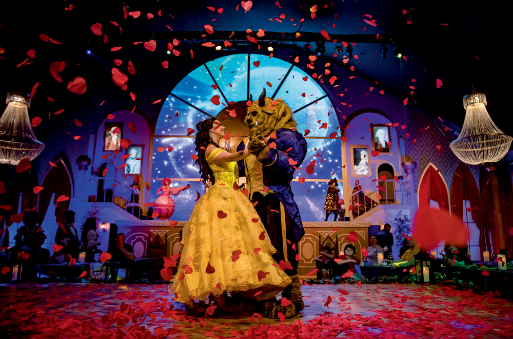 A imagem mostra a atriz da Bela e da Fera dançando no palco em uma chuva de pétalas de rosa.