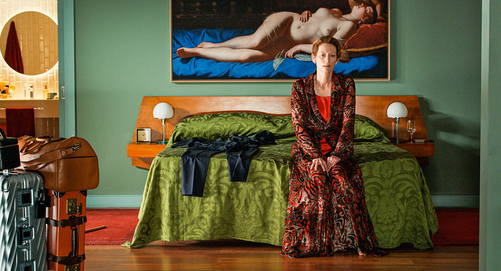 A imagem mostra Tilda Swinton, sentada em uma cama, olhando com rosto de desolação para frente.
