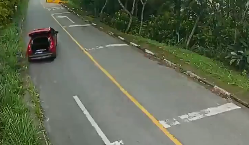 A imagem mostra um carro vermelho, em uma estrada, com o porta-malas aberto e em movimento.